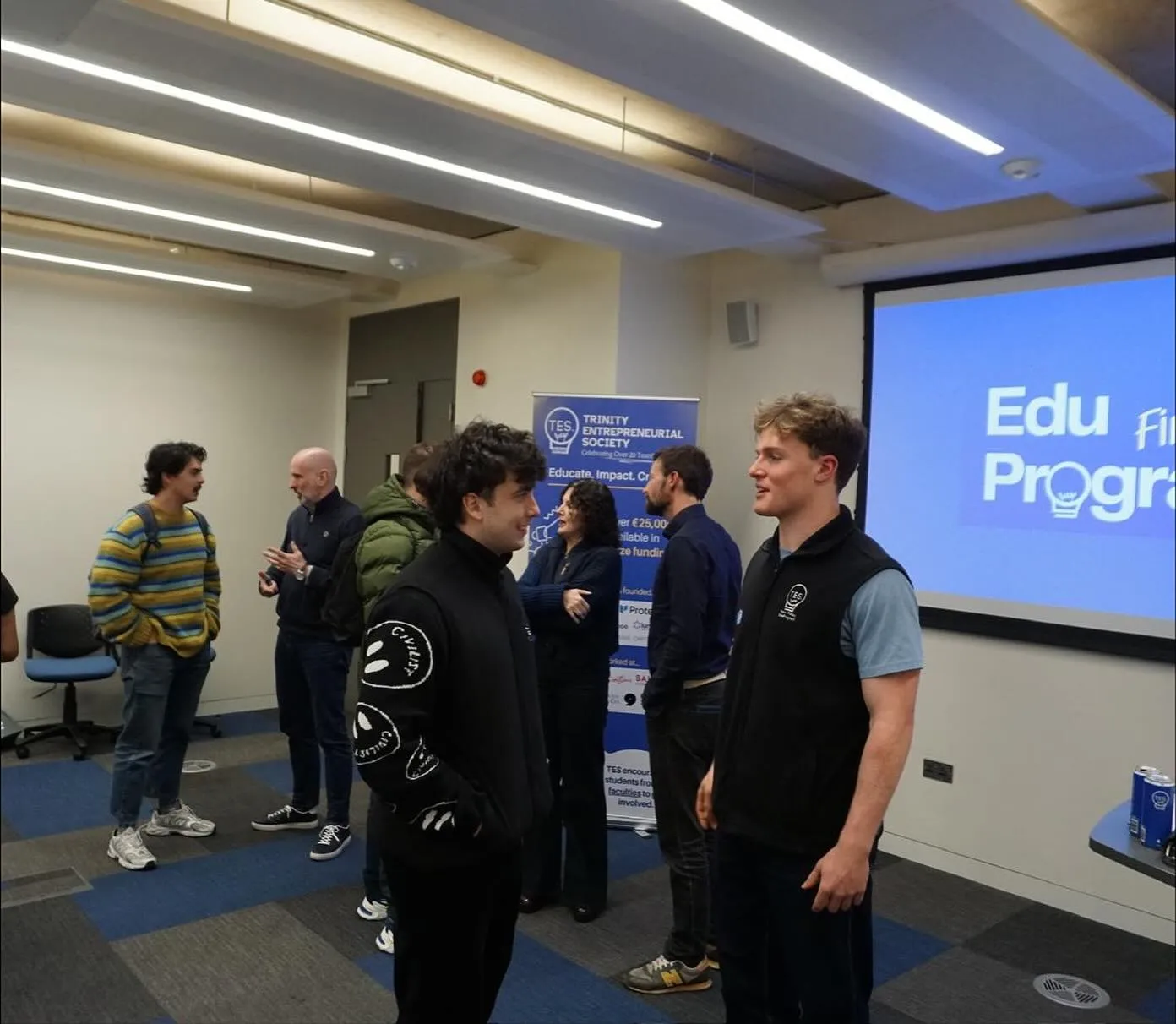 Students and speakers talking after an EduProgram session at Trinity Business School, beside a Trinity Entrepreneurial Society banner.